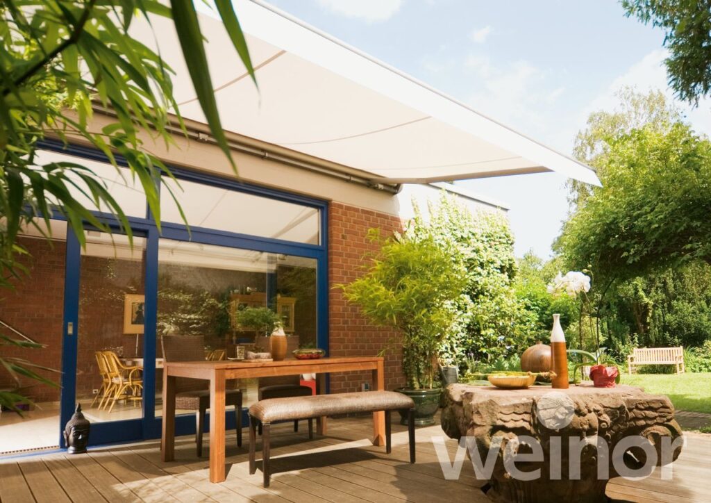 Sonnige Gartenterrasse mit einer beige Topas Markise von Weinor, ohne Dach, über einem gemütlichen Essbereich, eingebettet in üppiges Grün und einen gepflegten Garten, sichtbar durch große Fenstertüren eines roten Backsteinhauses.