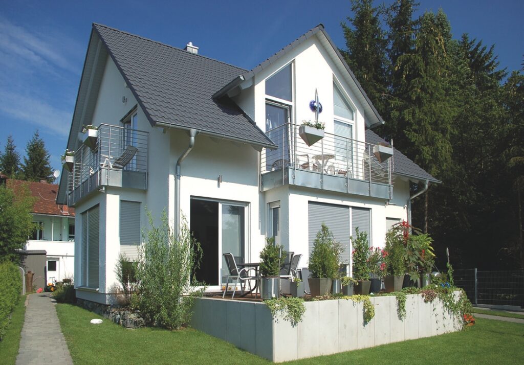 Modernes zweistöckiges Haus mit schiefergrauem Dach und weißen Außenwänden. Das Haus verfügt über große Fenster mit Rollläden, Balkone mit Edelstahlgeländern und eine Terrasse mit Gartenmöbeln. Der Vorgarten ist gepflegt mit Pflanzen in Töpfen und einem Rasen, umgeben von einem Betonzaun. Im Hintergrund sind hohe Bäume und ein benachbartes Haus zu sehen.